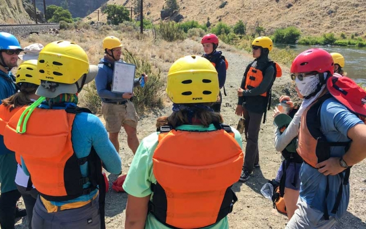 a group of students wearing life jackets and helmets watch as an instructor gives a rafting lesson